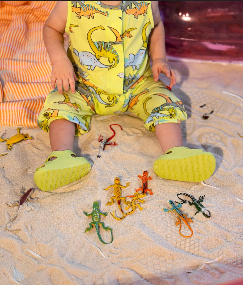 Child wearing a yellow outfit with dinosaur patterns and green shoes, standing on sand with toy lizards.