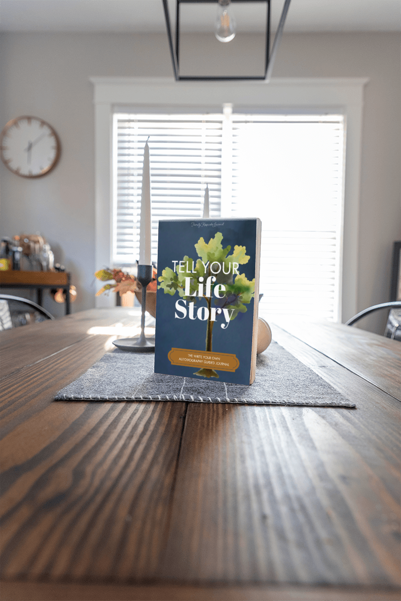 Book titled 'Tell Your Life Story' on a wooden table in a room with a clock and window blinds.