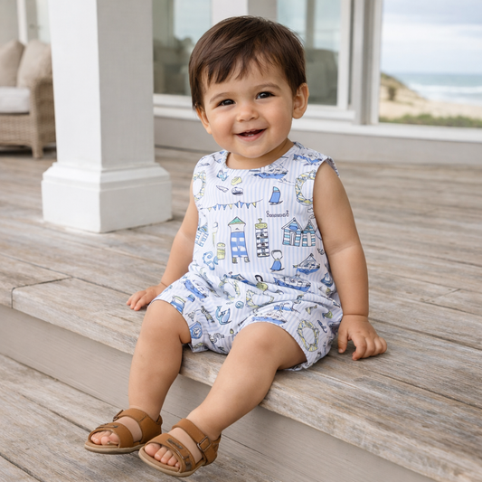 Child sitting on a wooden deck wearing a patterned romper with a coastal design.