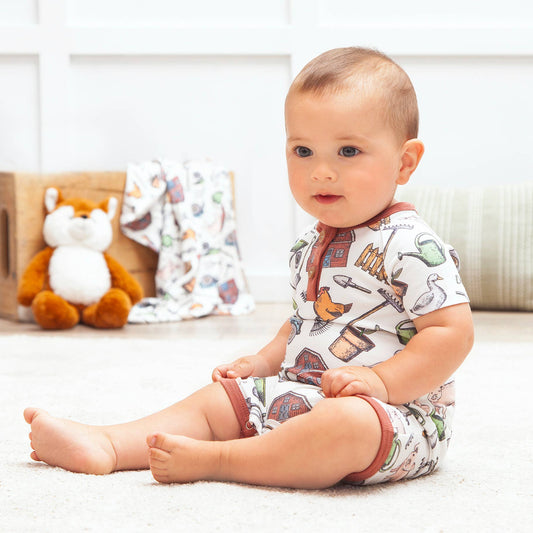 Baby sitting on a white surface wearing a patterned onesie with toys in the background.