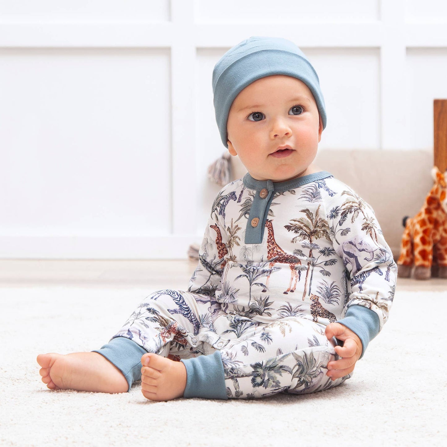 Baby wearing a patterned onesie and blue hat sitting on a white surface.