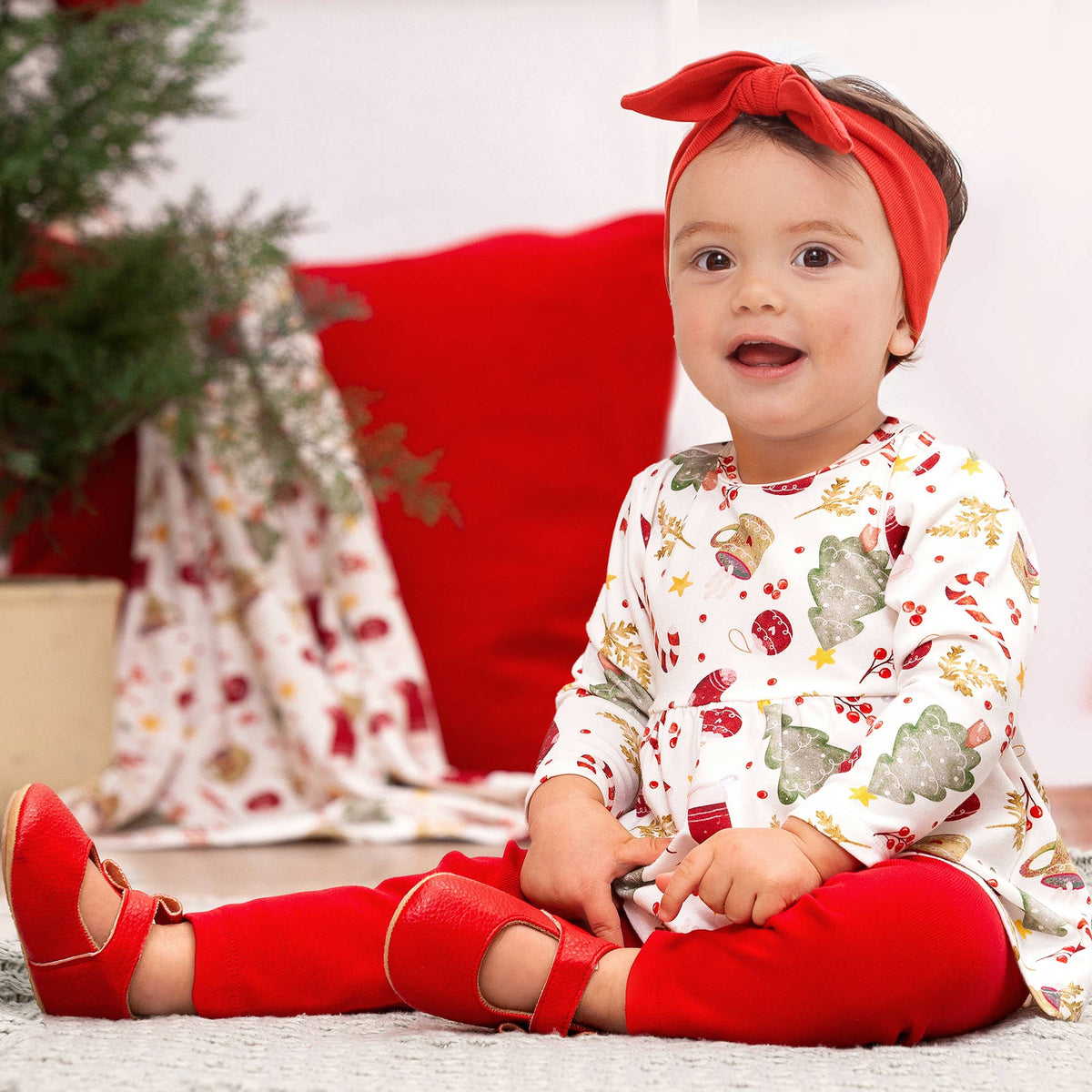 A baby girl  sitting in a room decorated for Christmas, wearing clothing that includes a Christmas-themed bamboo top and red leggings.