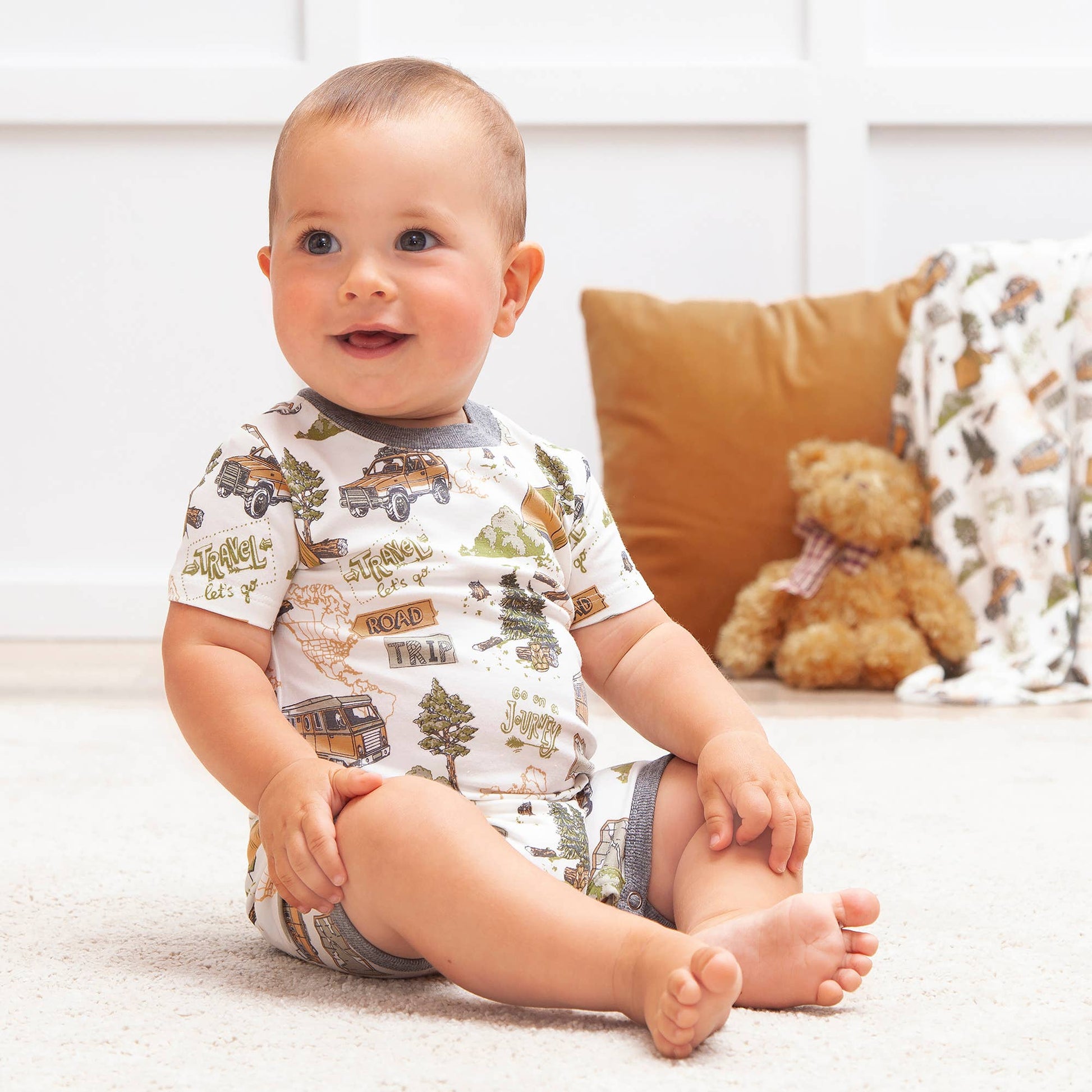 Baby sitting on a carpeted floor wearing a patterned onesie with a teddy bear and pillow in the background.