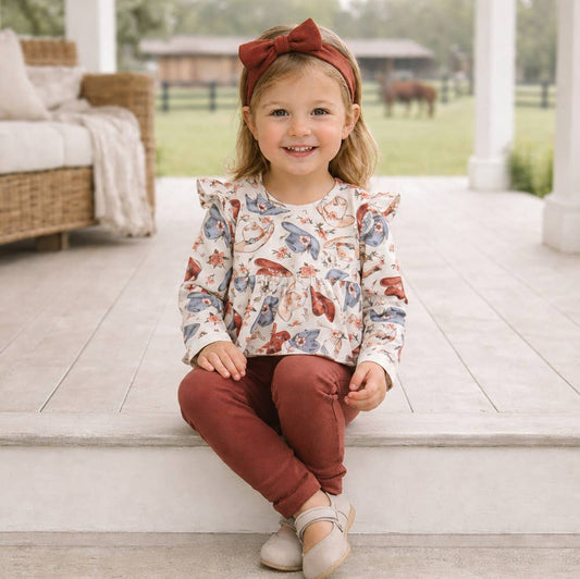 A young girl with a brown bow in her hair sits on white porch steps wearing a long-sleeve ruffled blouse with a colorful cowgirl hat pattern, brown colored pants, and beige shoes.