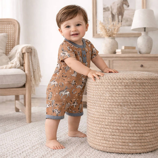 Baby in a western patterned romper standing next to a woven ottoman in a home setting.