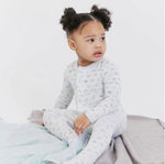 Child wearing a light blue long-sleeve one piece with an airplane pattern, sitting on a bed.