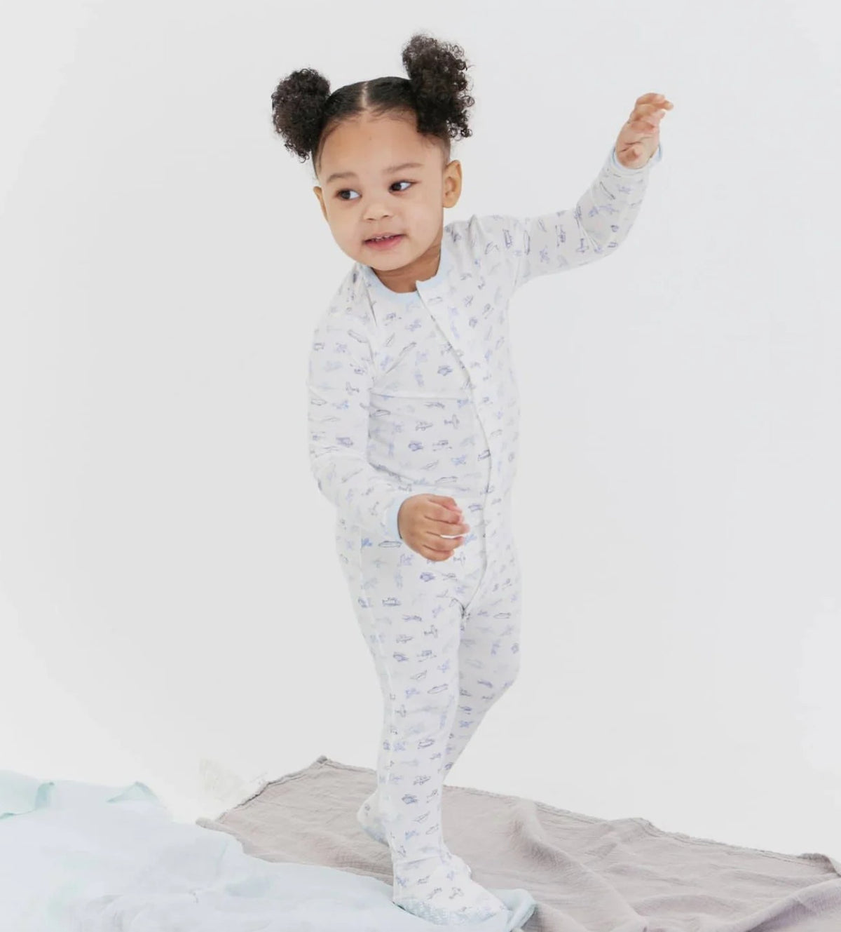 Child in a light blue onesie with a airplane pattern, standing on a bed with a white background