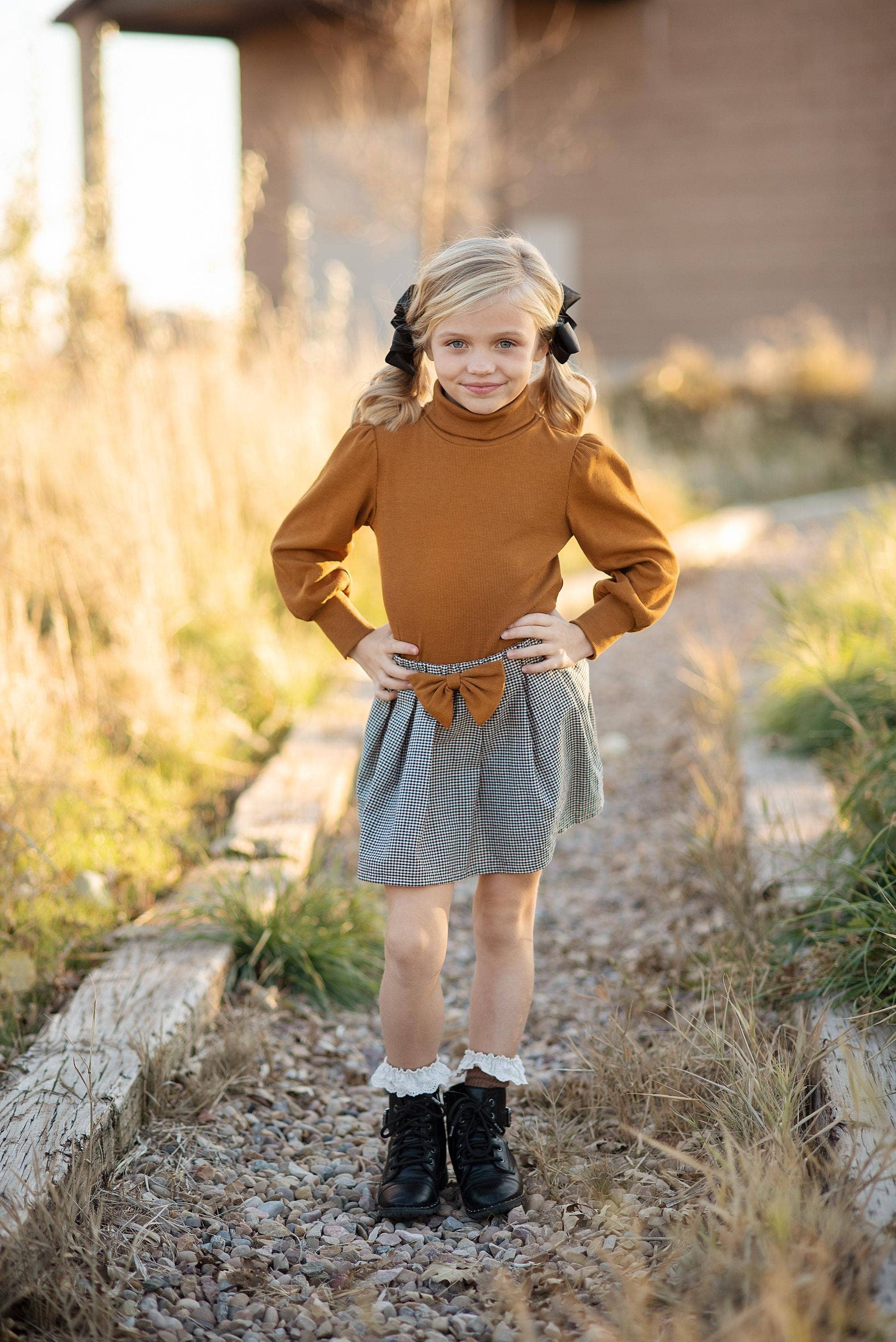 A girl wearing a caramel colored long puff sleeve top with a black and white houndstooth check pleated skirt with matching bow, standing on a gravel path.