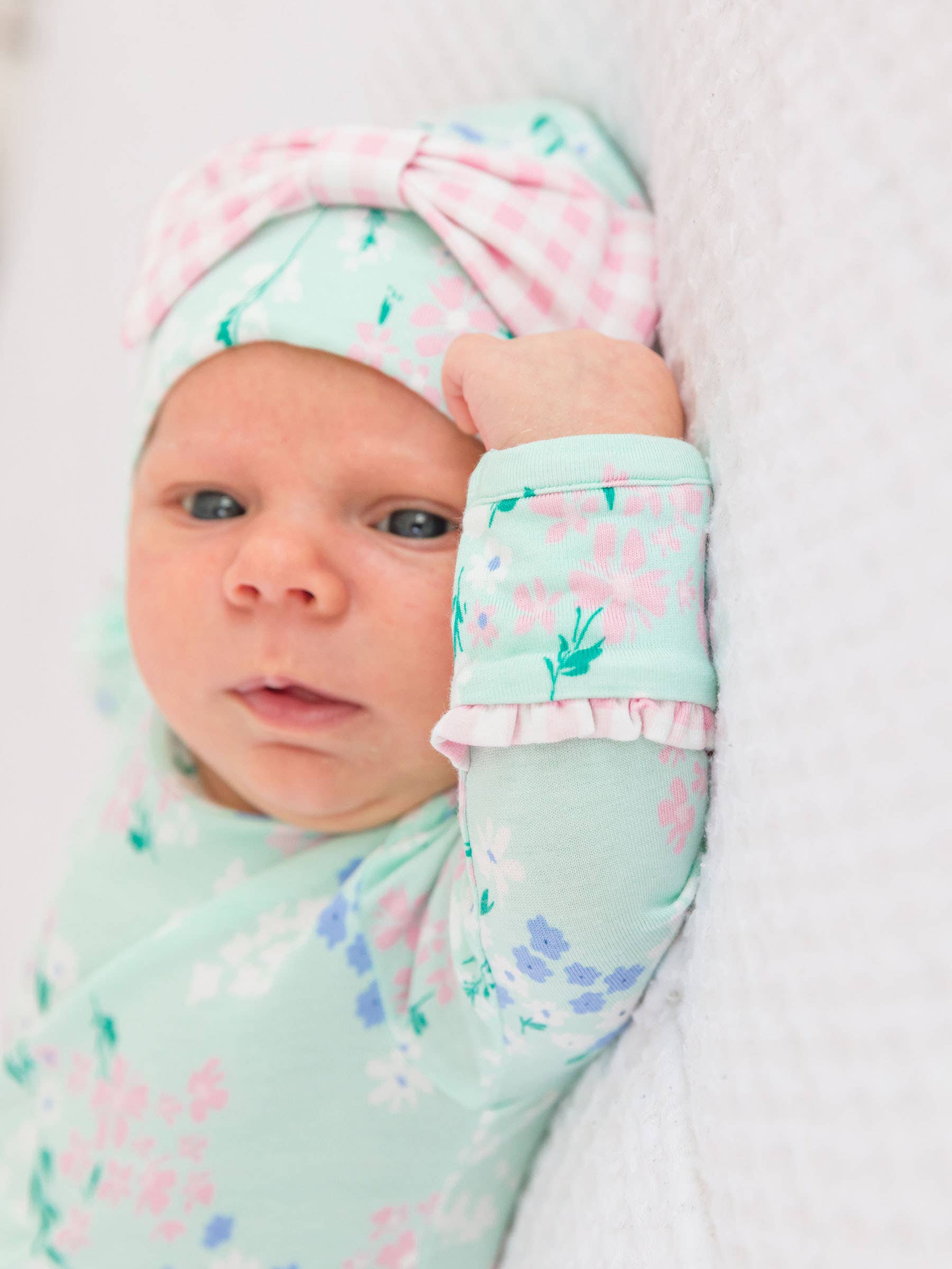 Newborn baby wearing a floral beanie hat and matching knotted gown on a white background