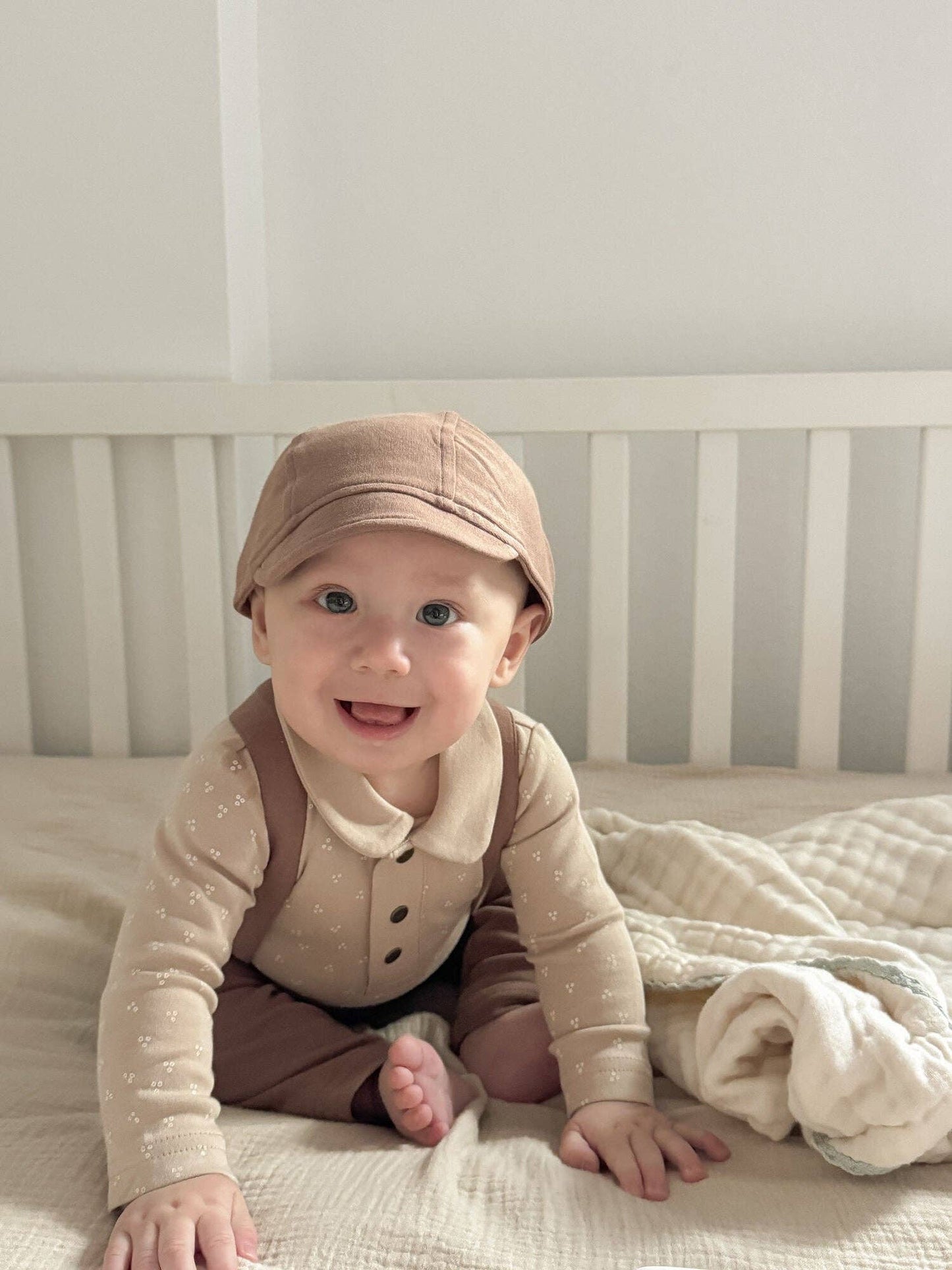 Baby sitting in a crib wearing a brown cap and outfit