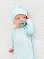 Baby in a light blue checkered outfit with matching hat and pacifier on a white textured blanket.