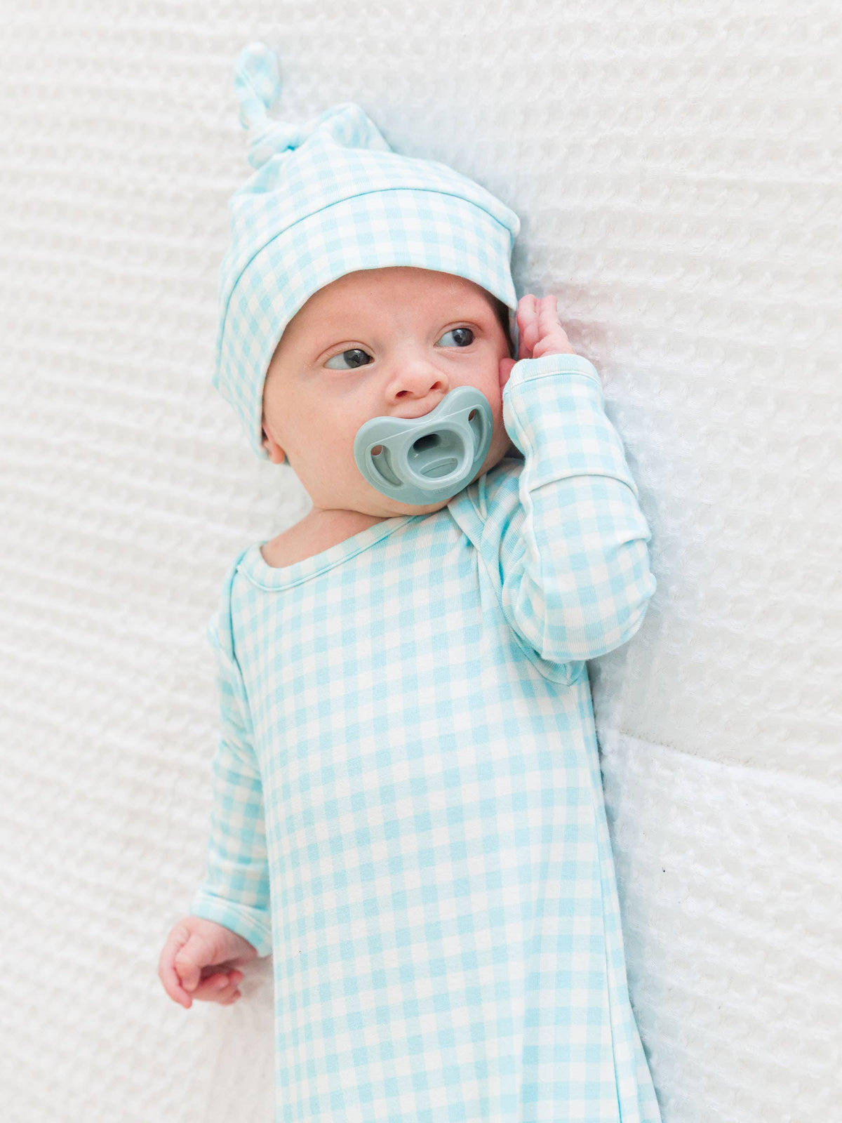 Baby in a light blue checkered outfit with matching hat and pacifier on a white textured blanket.