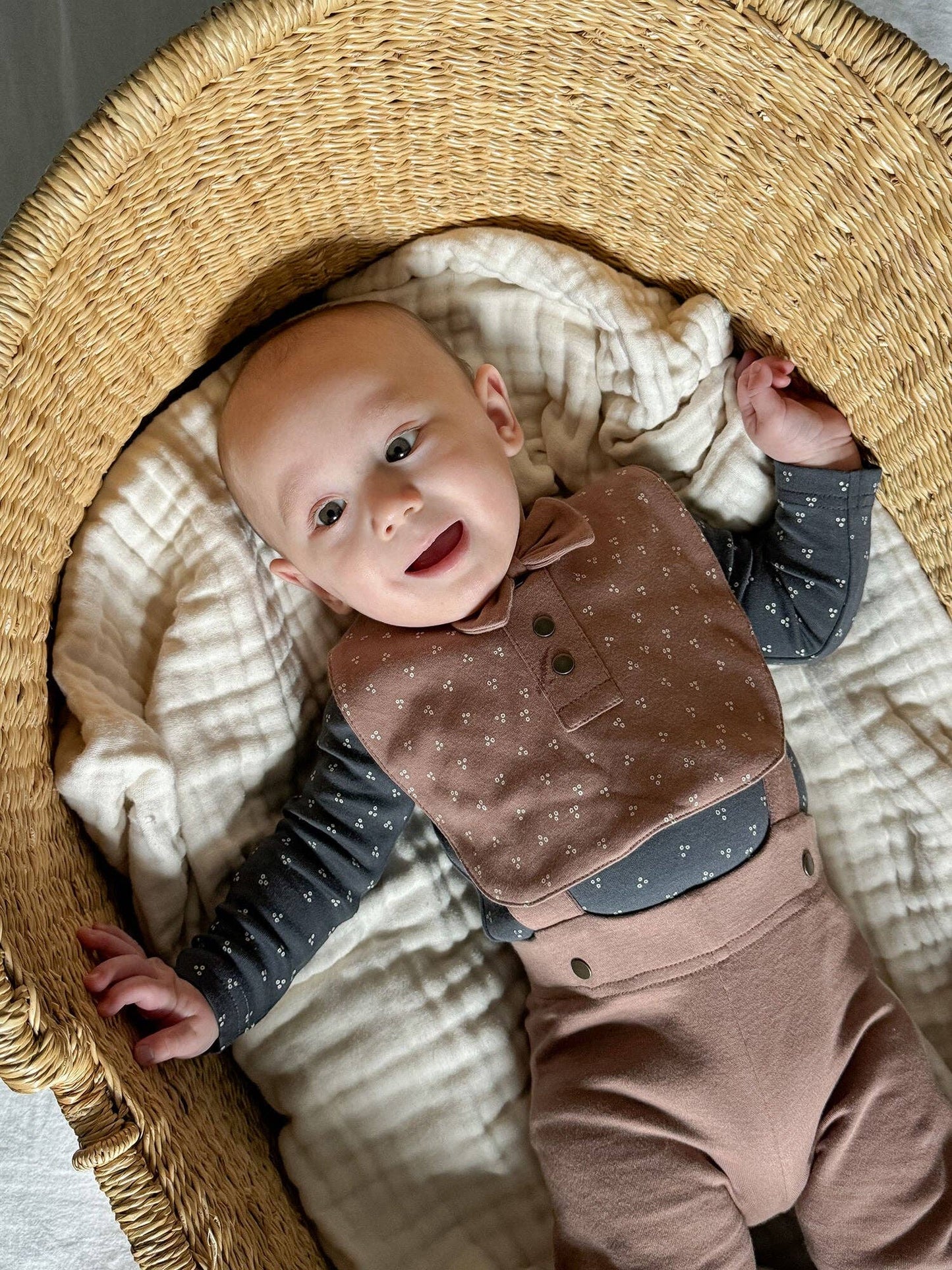 Baby in a woven basket wearing a brown bib and pants with charcoal shirt.