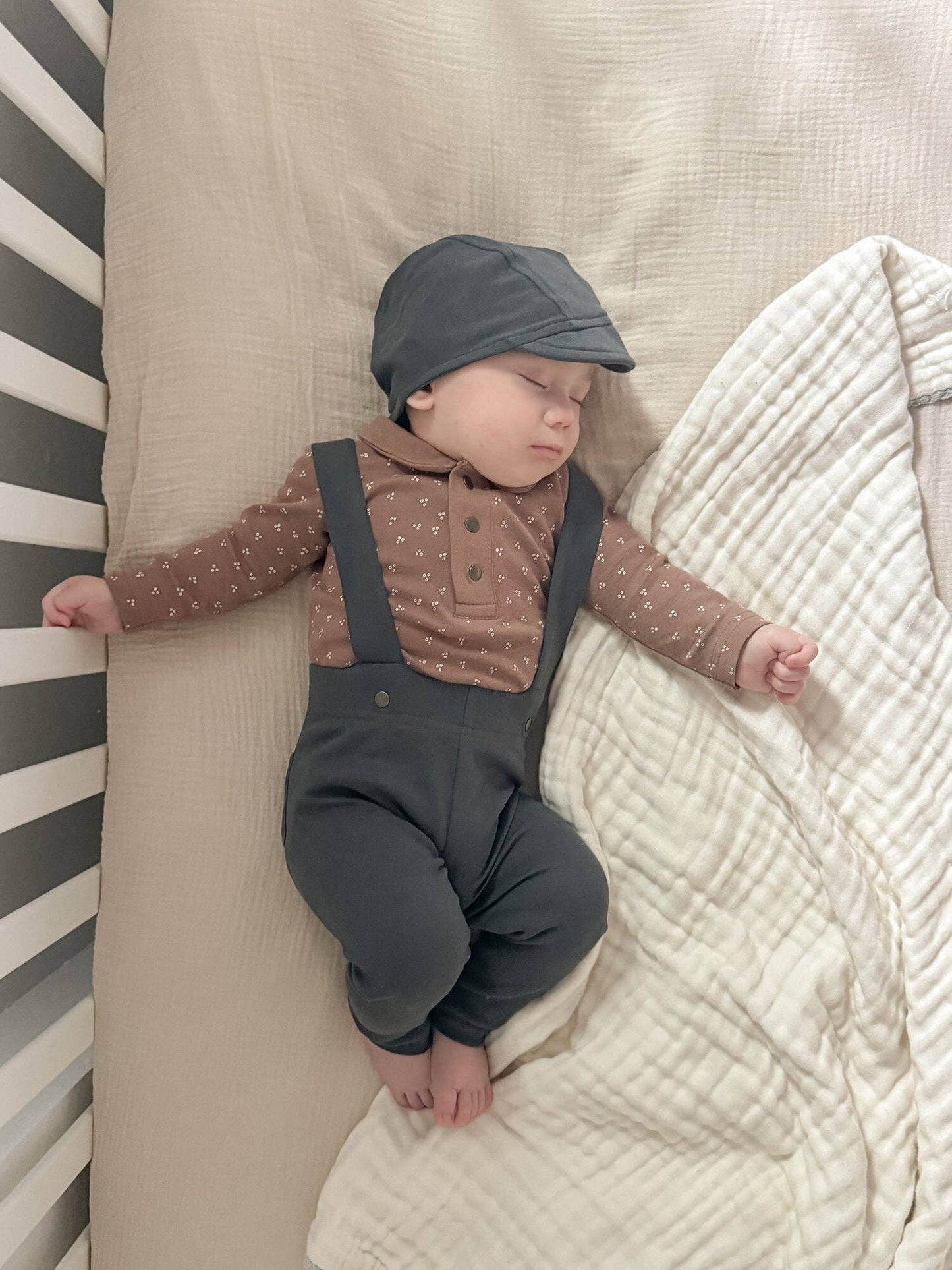Baby in a brown patterned shirt and dark pants with suspenders lying on a textured surface.