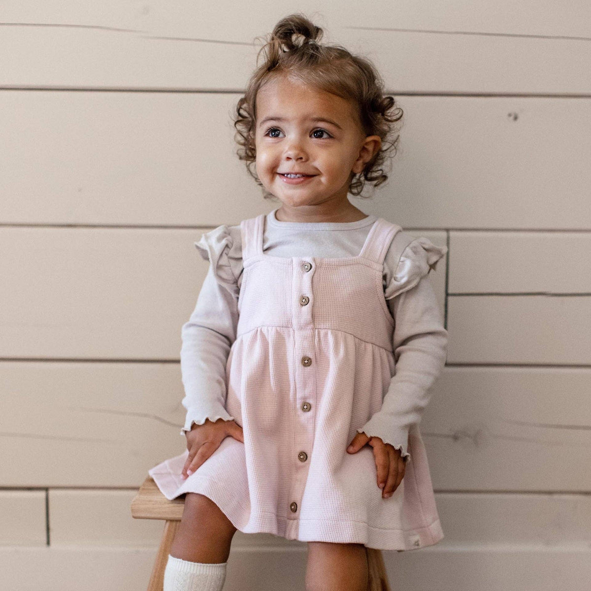 Young girl wearing a pink dress with a beige shirt, sitting on a wooden stool against a wooden wall.