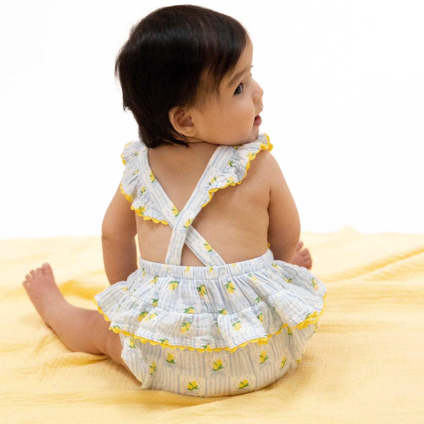 back of Baby wearing a white romper with yellow accents on a yellow blanket