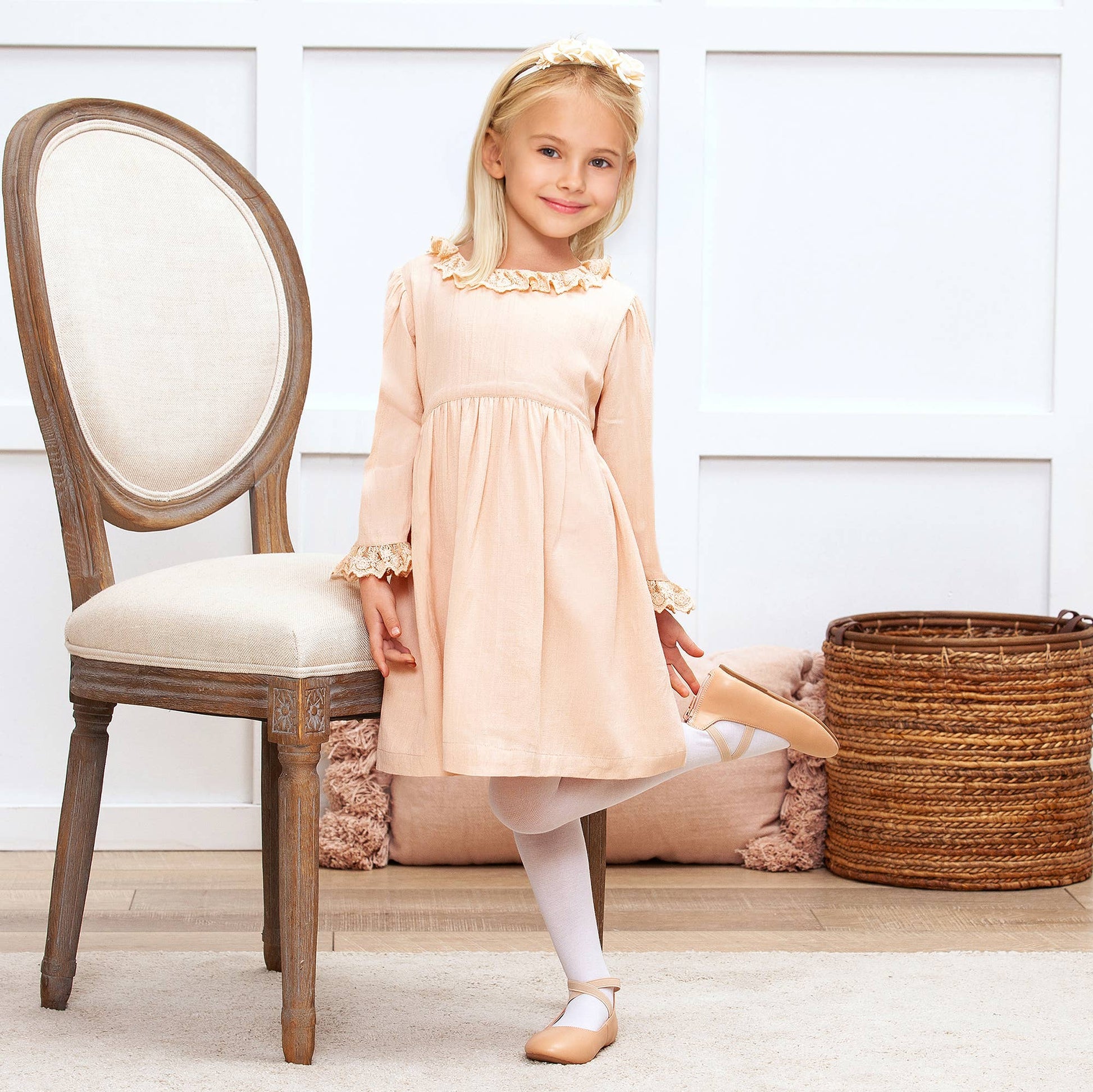 Young girl in a beige dress standing next to a wooden chair in a room with a white wall and wooden floor.