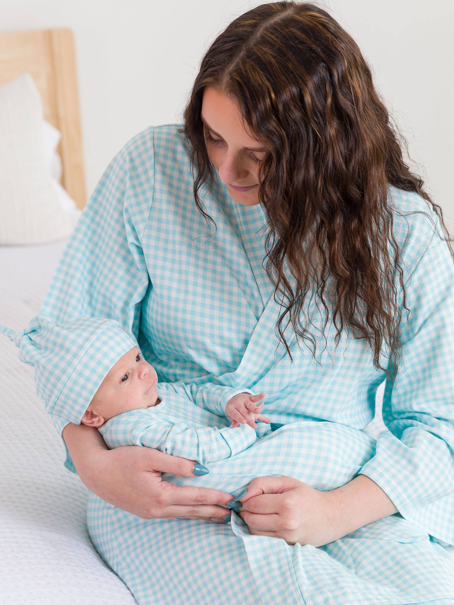 Woman in a light blue checkered robe holding a baby in a matching outfit on a white bed.