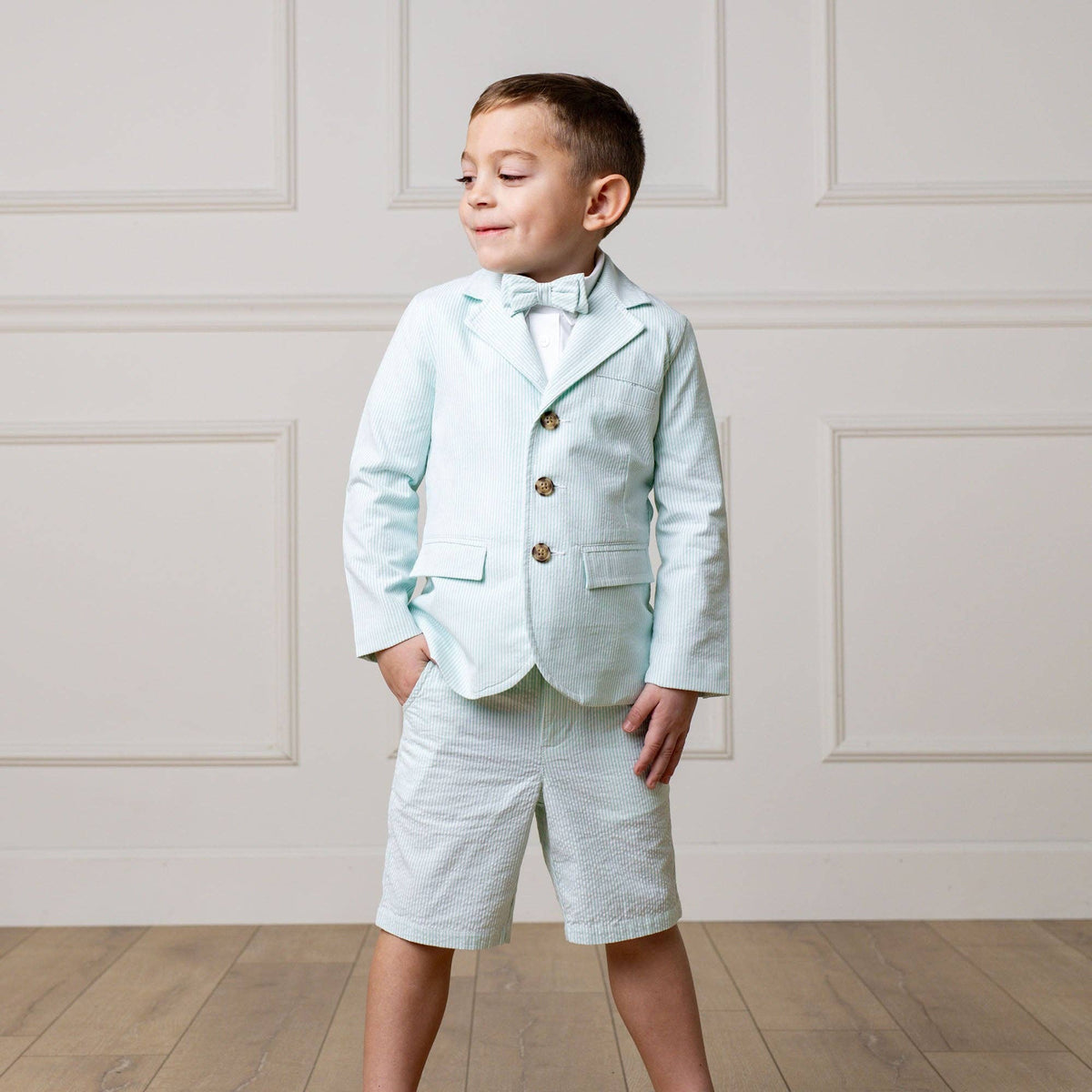 Young boy in a mint suit with a bow tie standing against a white paneled wall.