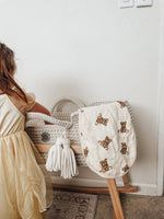 Woman in a light dress standing next to a baby crib with a white burp cloth featuring brown teddy bear patterns.