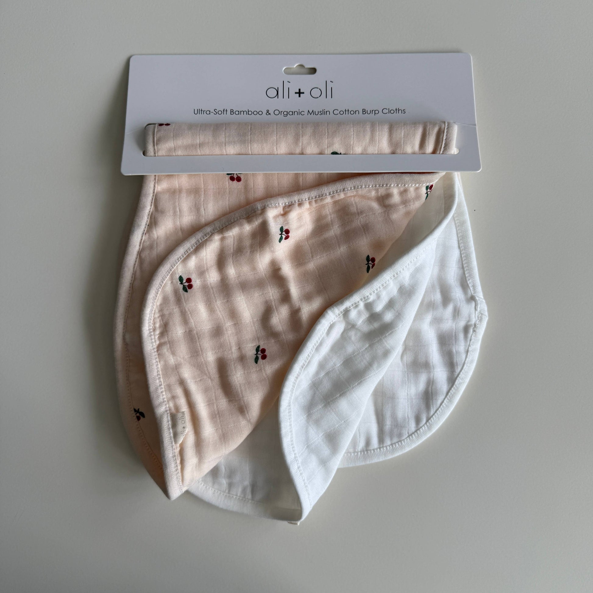 Two baby burp cloths, one pink with cherry pattern and one white, on a light gray background with 'ali+oli' branding.