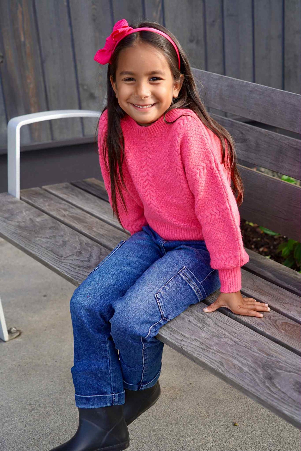 Young girl in a pink sweater and blue jeans sitting on a wooden bench.