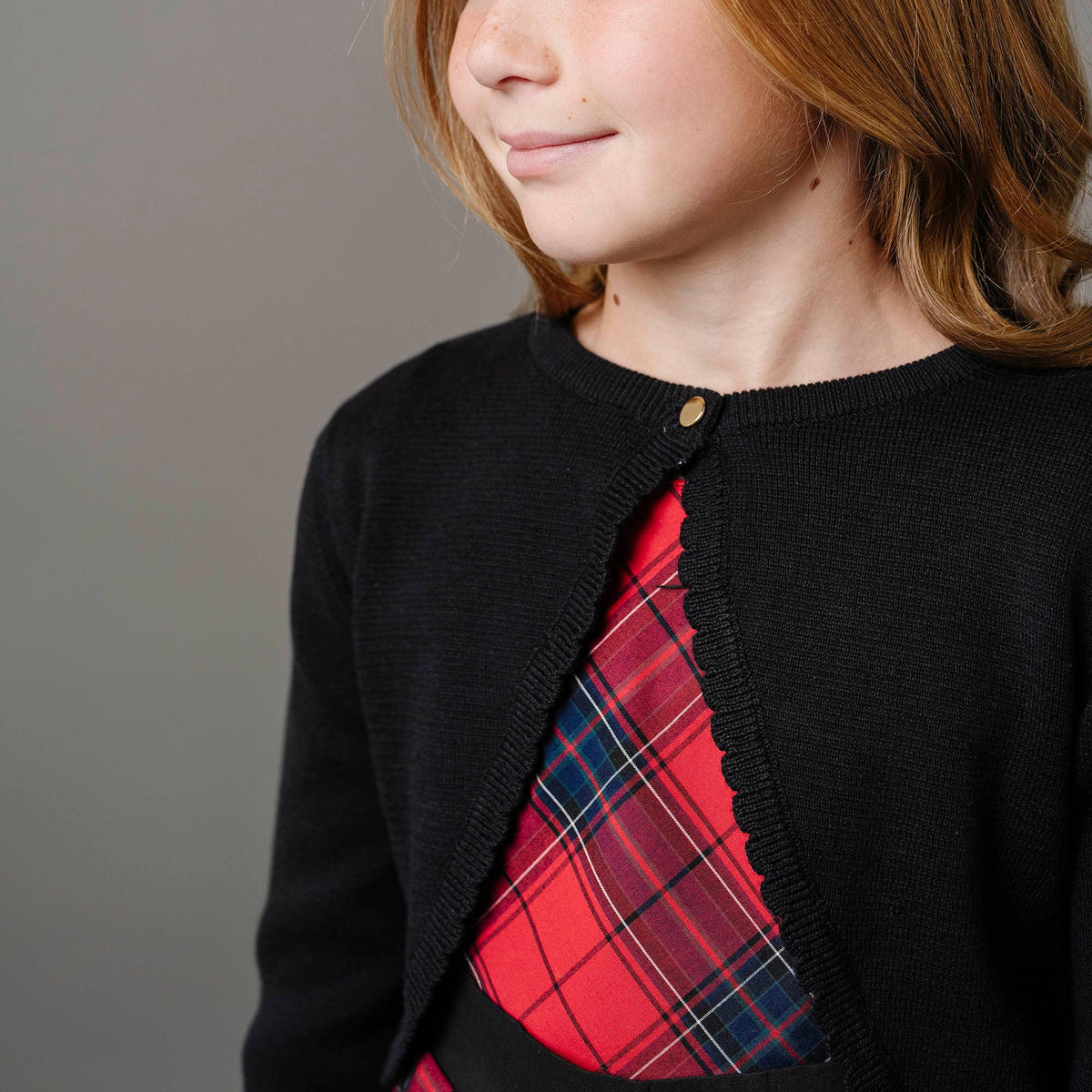 Child wearing a black cardigan over a red plaid shirt on a gray background