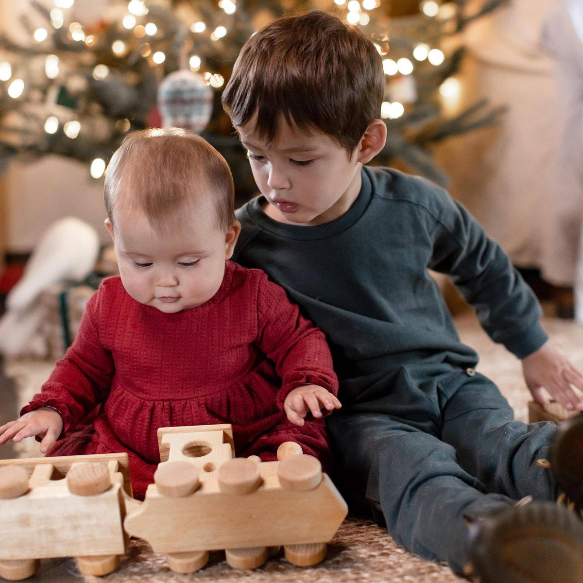 Two children playing with wooden toys in front of a decorated Christmas tree.