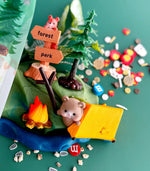 Miniature scene with a mouse by a campfire and tent, surrounded by colorful elements on a green background.