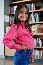 Young girl in a pink sweater standing in front of a bookshelf