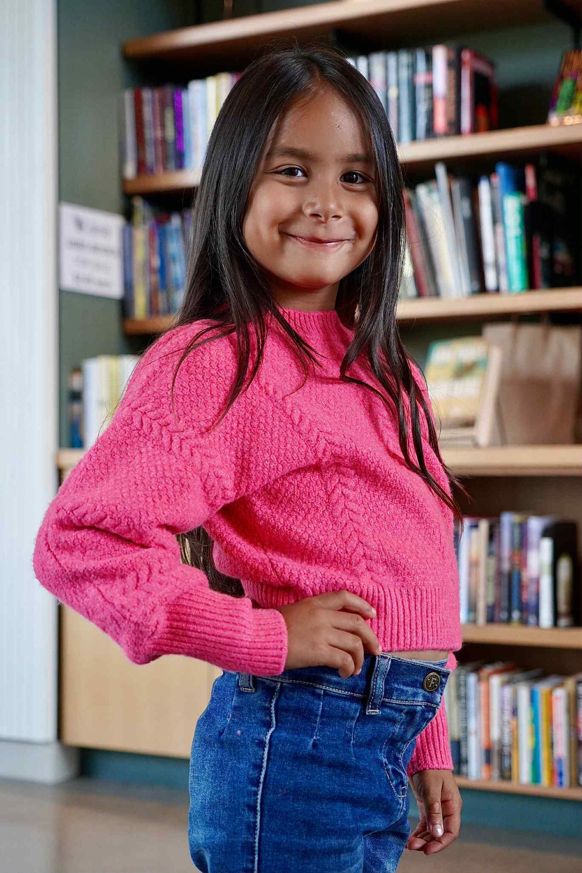 Young girl in a pink sweater standing in front of a bookshelf