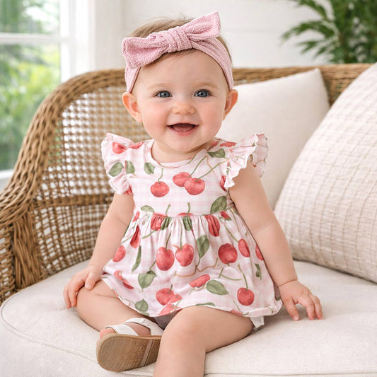 Baby wearing a cherry print dress and pink headband sitting on a couch.