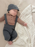 Baby wearing a brown shirt and charcoal pants and cap, sleeping on a beige blanket.