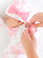 Baby in pink and white toile outfit with a large bow, being held by a person.