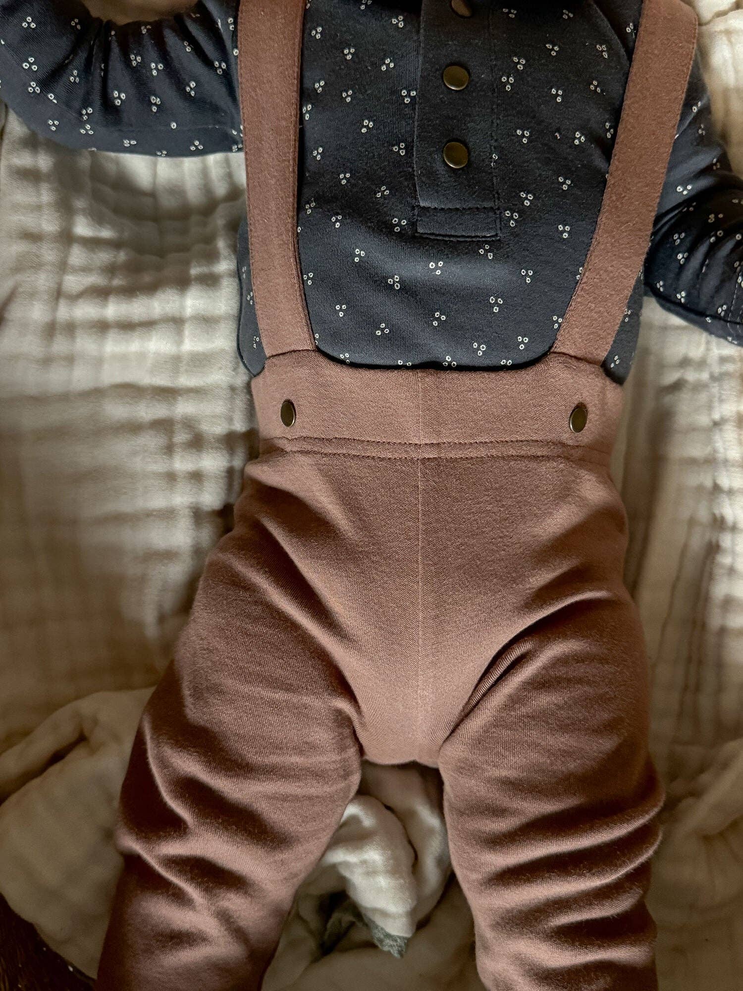 Brown pants worn by a baby on a bed with a patterned shirt underneath.