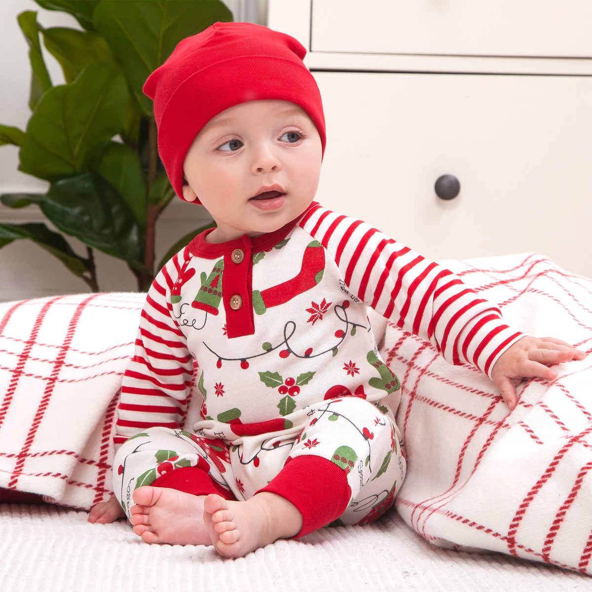 Baby in red and white striped outfit with holiday pattern, sitting on a plaid blanket.