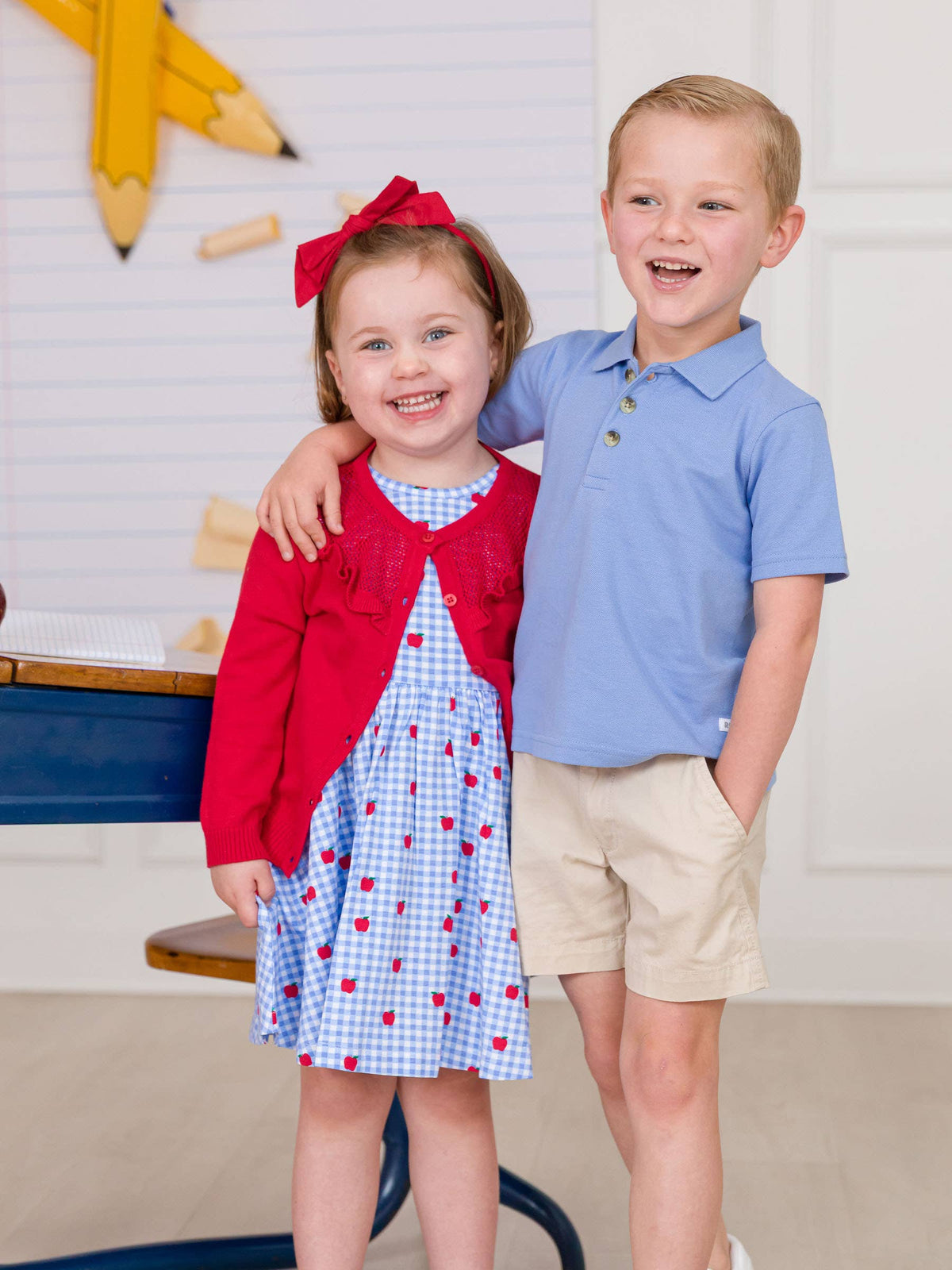 Two children, a girl in a red cardigan and checkered dress, and a boy in a blue shirt and beige shorts, standing together indoors.