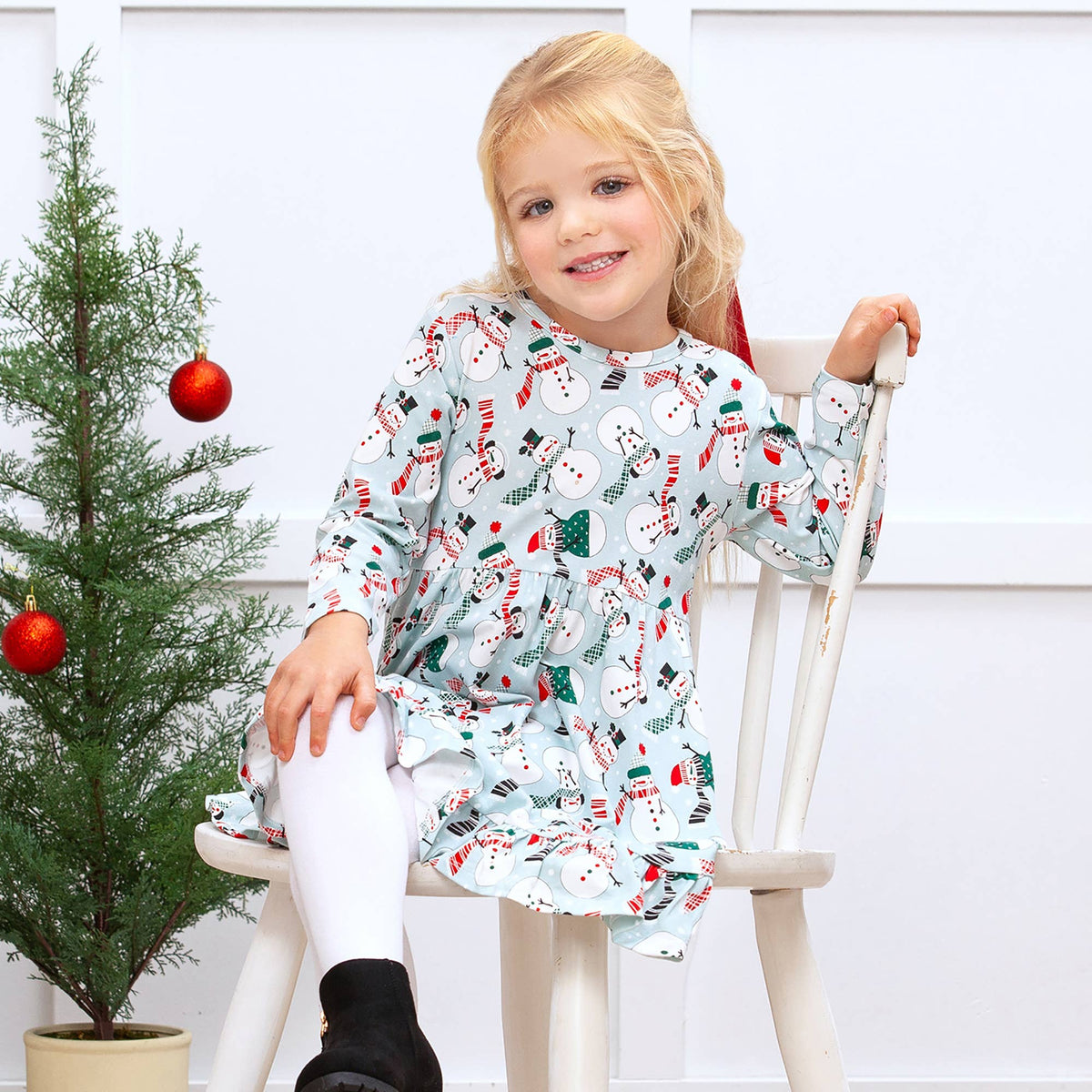 Young girl in a festive dress sitting on a chair next to a small Christmas tree.