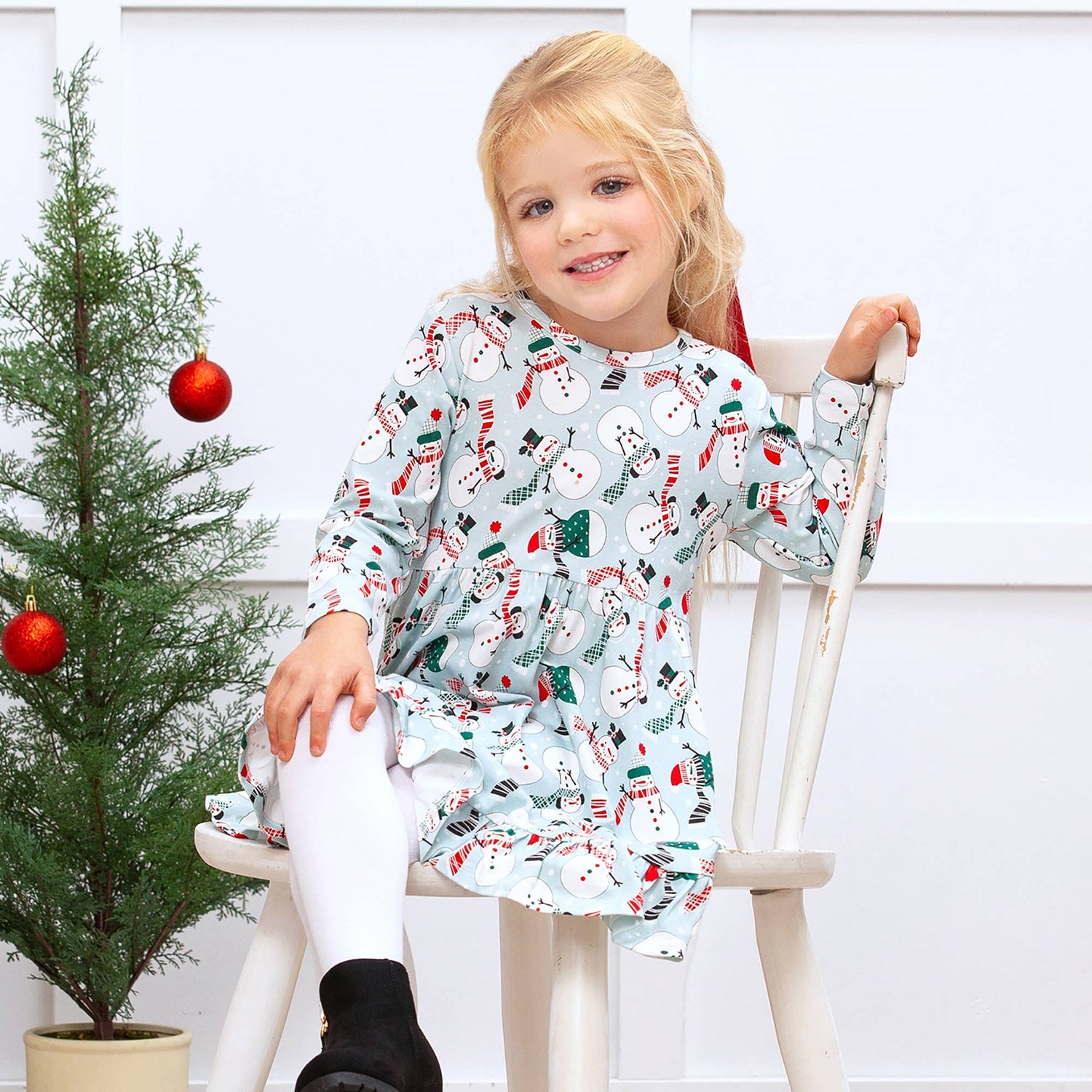 Young girl in a festive dress sitting on a chair next to a small Christmas tree.