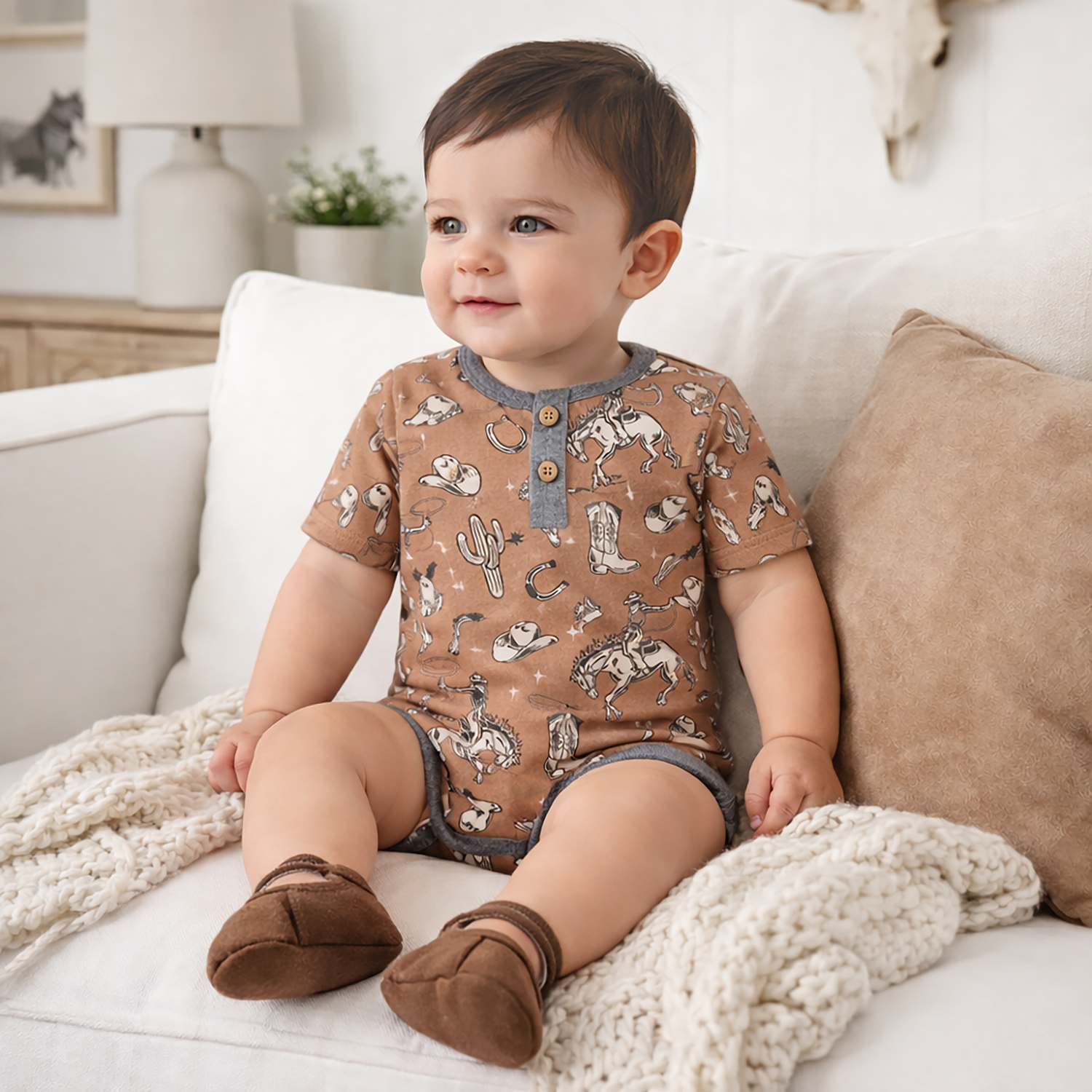 Baby wearing a brown romper with cowboy print sitting on a couch.