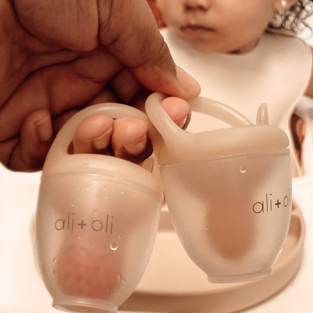 Two ali+oli baby cups held by a hand with a child in the background.