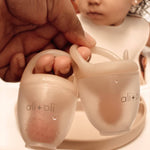 Two ali+oli baby cups held by a hand with a child in the background.
