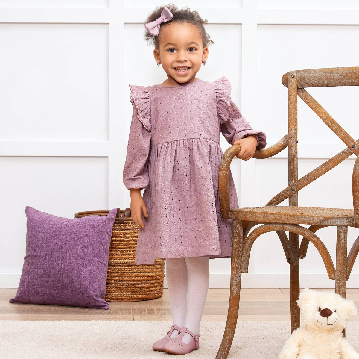 Young girl in a mauve eyelet dress standing next to a wooden chair and purple pillow indoors.