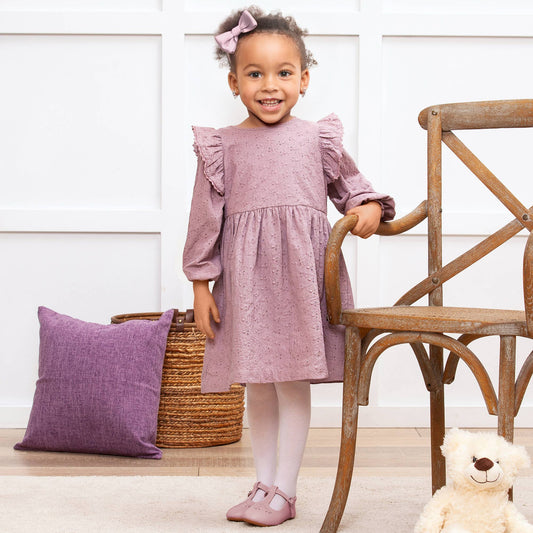 Young girl in a mauve eyelet dress standing next to a wooden chair and purple pillow indoors.