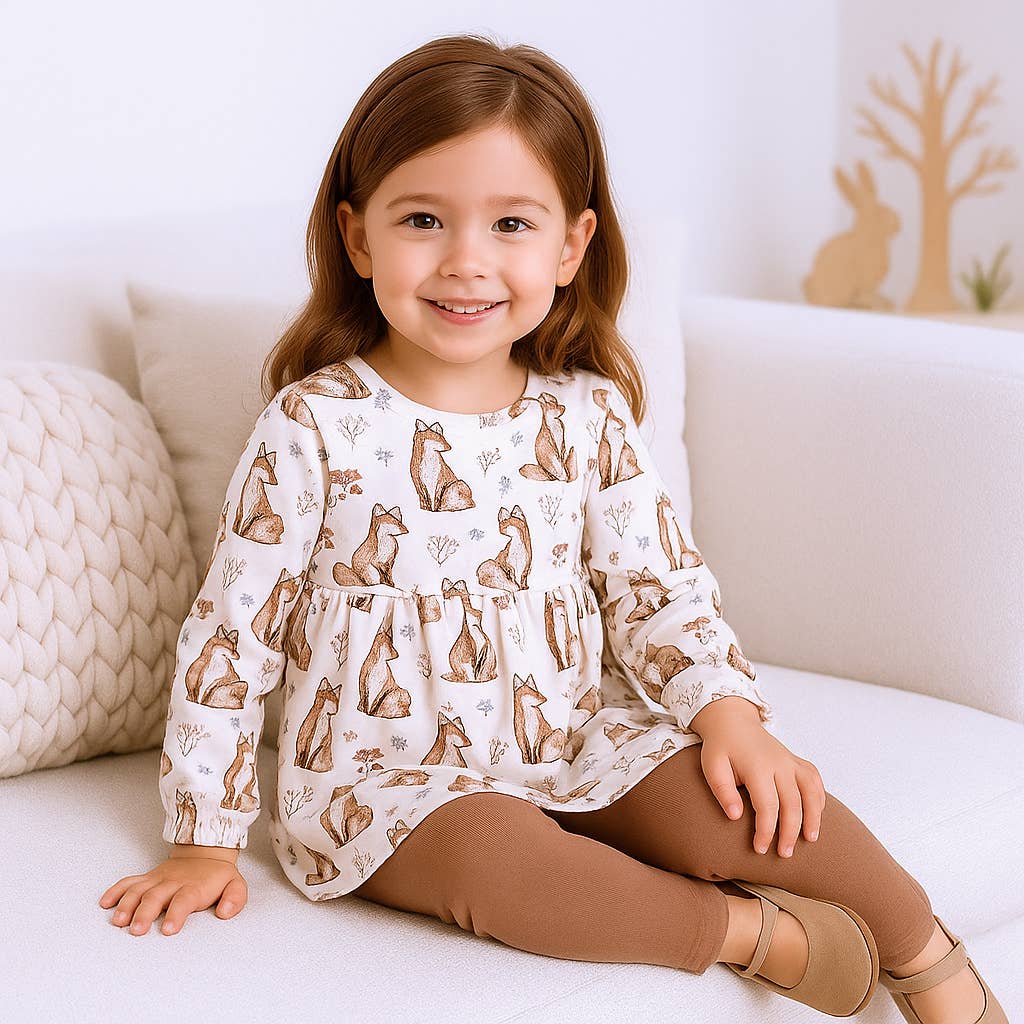 Young girl wearing a white long-sleeve shirt with fox patterns, sitting on a white couch.