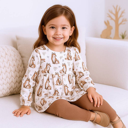 Young girl wearing a white long-sleeve shirt with fox patterns, sitting on a white couch.