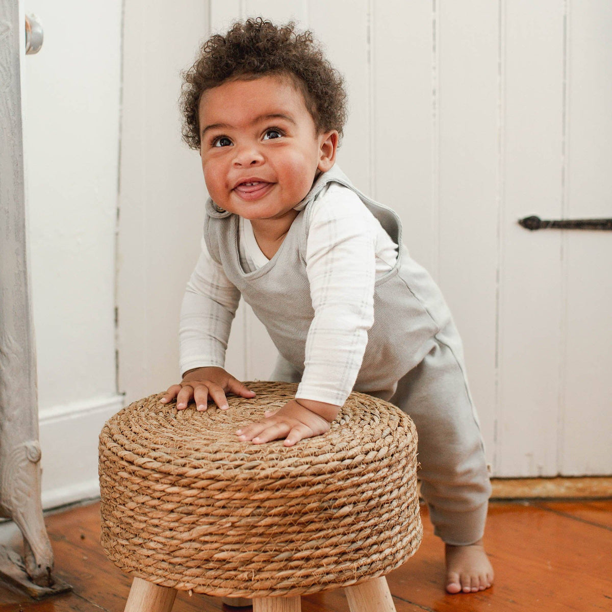 Child standing on a woven stool wearing a gray overall with white bodysuit