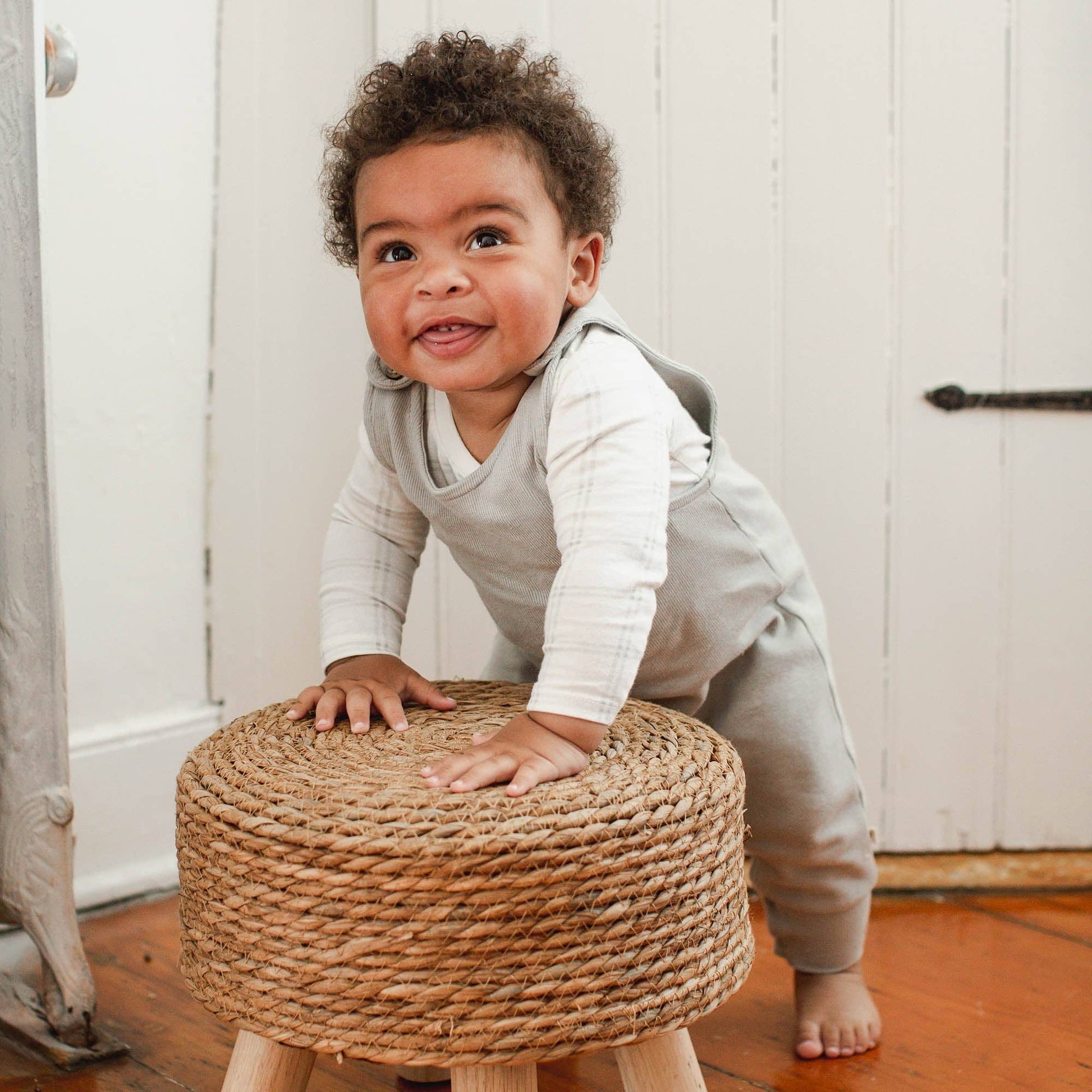 Child standing on a woven stool wearing a gray overall with white bodysuit