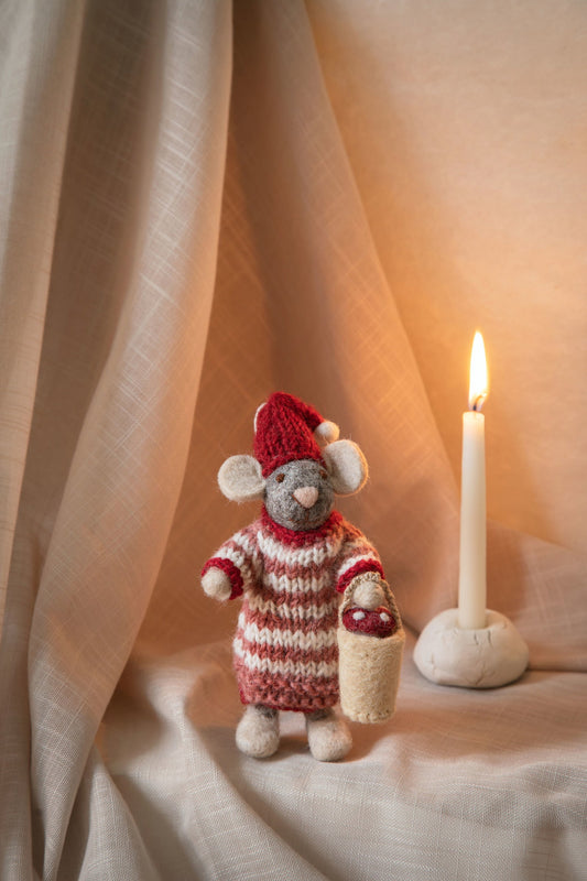 A small knitted mouse figurine wearing a red Santa hat and red-and-white striped sweater, holding a small basket, posed beside a lit white candle on draped beige fabric.
