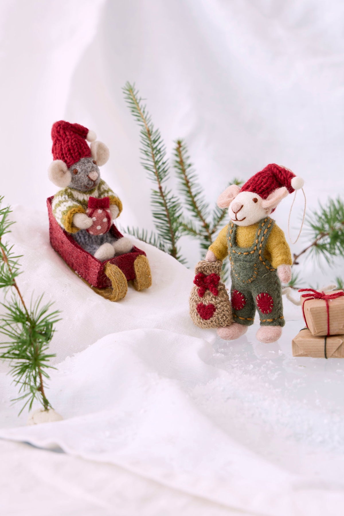 Handmade felt Christmas mouse ornaments styled in a snowy scene. One ornament depicts a gray mouse wearing a red knit hat and sweater, sitting in a red sled.